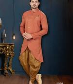 Coral-peach sherwani with textured finish, asymmetrical hem, pocket square, and gold trousers.