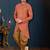Coral-peach sherwani with textured finish, asymmetrical hem, pocket square, and gold trousers.