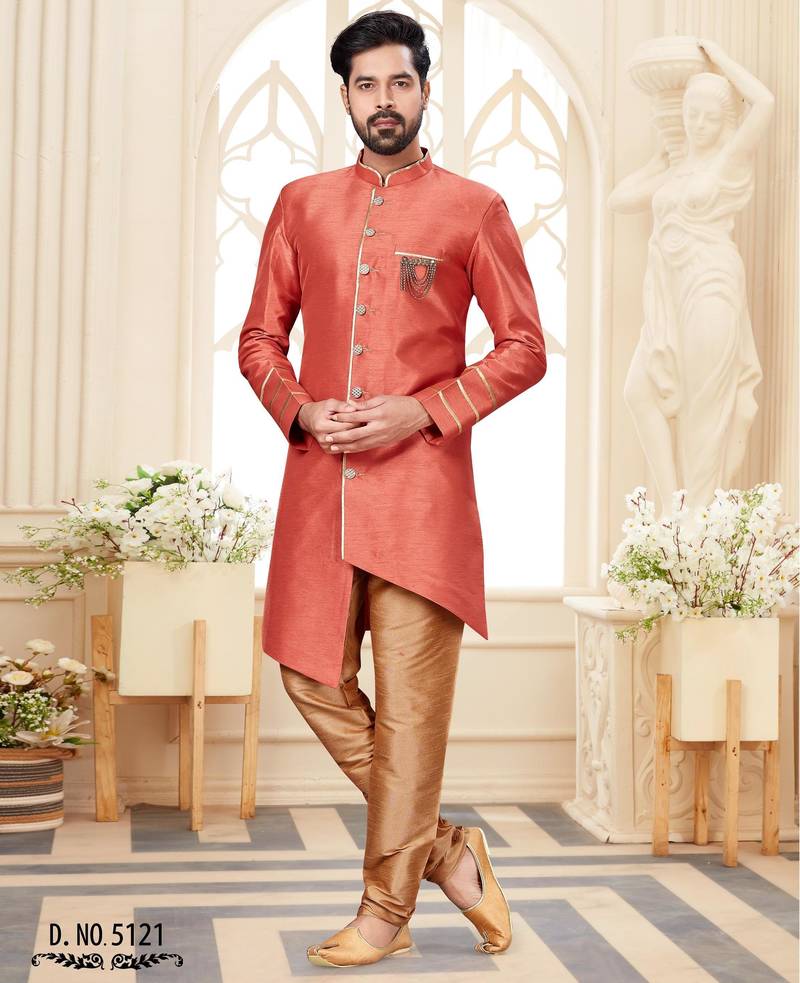 Coral-peach sherwani with gold piping, chain pocket, and gold trousers.